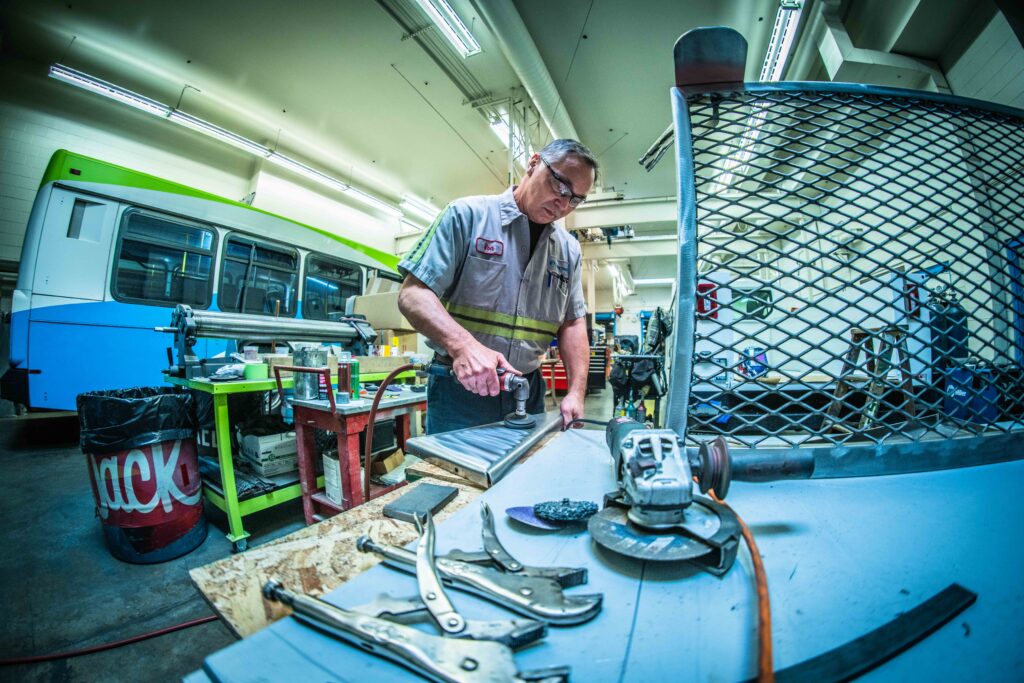 A man in a workshop uses a tool on a metal piece at a workbench. Various tools and equipment are spread out on the bench, and a bus is visible in the background. The workshop is well-lit with fluorescent lights, creating an ideal setting for job listings related to skilled trades.