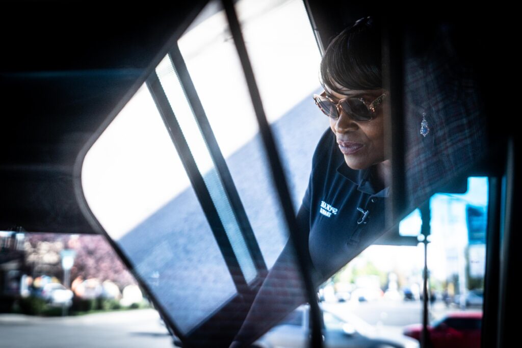 A person wearing sunglasses and a dark top labeled "DEBUG" is sitting inside a vehicle, looking out of a tinted window. The background includes blurred elements of an outdoor urban scene with buildings and signs. The interior reflections add a layered look to the image, reminiscent of retention in hiring bus drivers.