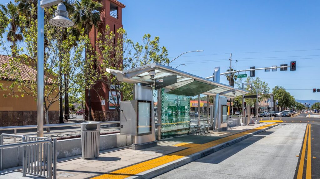 A modern, clean bus stop with a shaded seating area, trash cans, and an information board. The bus stop is situated on a sunny street lined with trees. Traffic lights and buildings are visible in the background. A bike lane runs parallel to the street.