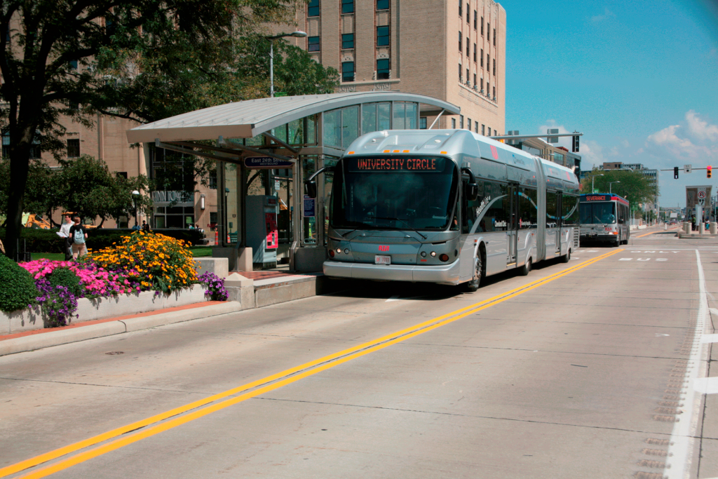 A silver bus with a "University Circle" sign on its front is stopped at a bus shelter on a sunny day. Colorful flowers adorn the landscaped area next to the shelter, and a tall building is visible in the background. Another bus is approaching in the distance.