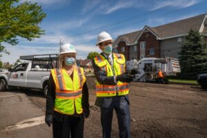 Two construction workers wearing white hard hats, protective masks, and high-visibility vests stand on a city line road under construction. The worker on the right points towards something off-camera. In the background, there's construction equipment and residential buildings.