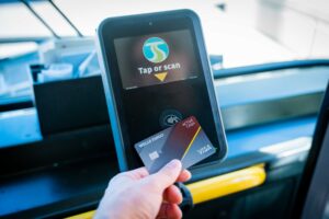 A hand holding a Wells Fargo Active Cash Visa credit card in front of a card reader on a bus. The card reader screen displays "Tap or scan" with an icon of a card being tapped.