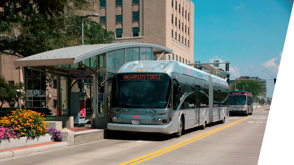 A modern city bus labeled "University Circle" is stopped at a bus stop in an urban area. The bus stop has a curved glass shelter, and there are flowers planted nearby. Another bus is visible in the background, and tall buildings surround the scene.