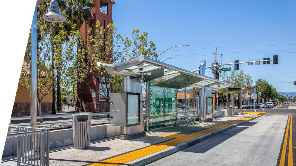 A modern bus stop with a glass shelter, metal bench seating, and tactile paving for accessibility. The stop is located on a sunny street with a few trees and a traffic light in the background. A red building with a tower is visible to the left.