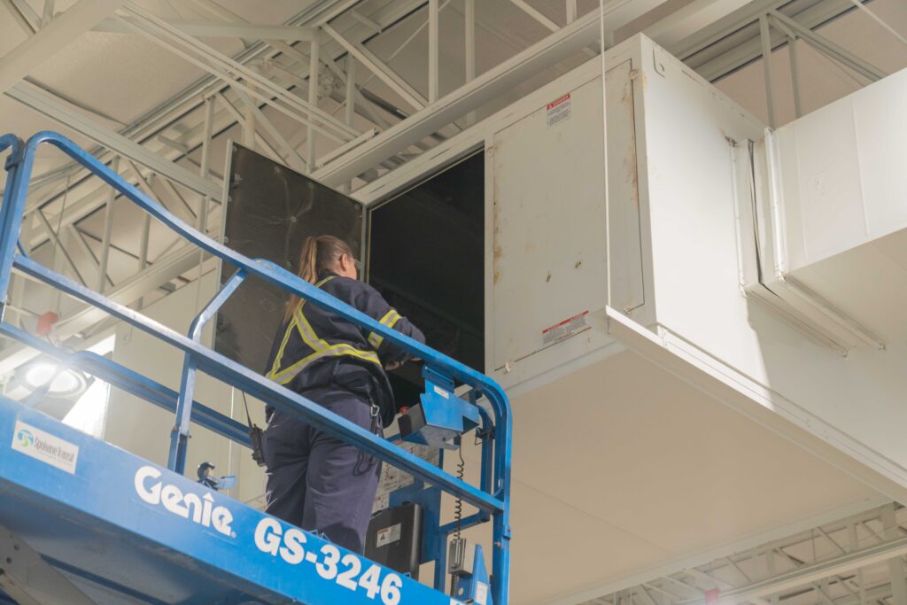 An STA-employed HVAC specialist on a lift inspects a large air duct under the high ceiling.