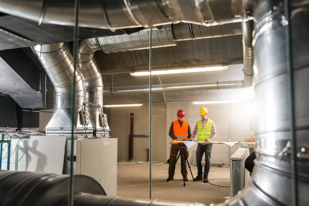 Two construction workers in hard hats and reflective vests consult a blueprint in an industrial setting with large metal ventilation ducts and unfinished walls. One worker holds a clipboard while the other inspects the plans under bright overhead lighting.