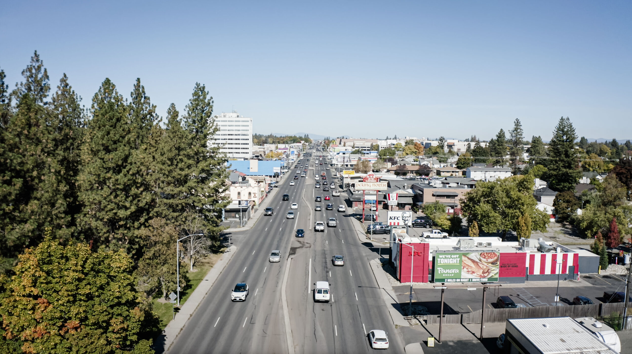 An aerial view of a busy multi-lane road lined with trees and various commercial buildings, including shops and restaurants. Several cars are driving in both directions, and the weather appears clear with a bright blue sky.