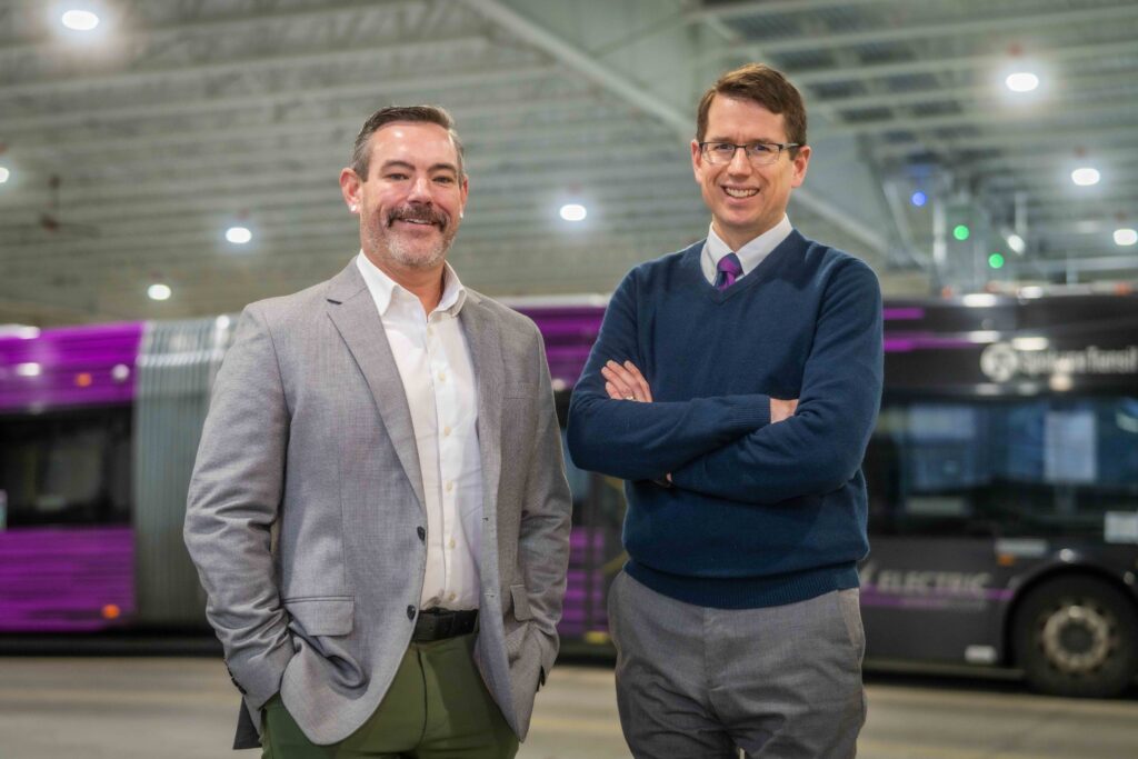 Two men standing in a bus depot, one in a suit and the other in a sweater, with purple buses in the background.