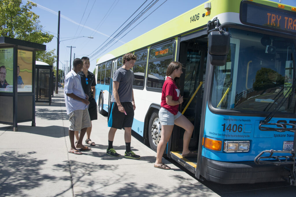 A group of people board a blue and green public bus at a bus stop on a sunny day, many clutching their STA Student Passes.