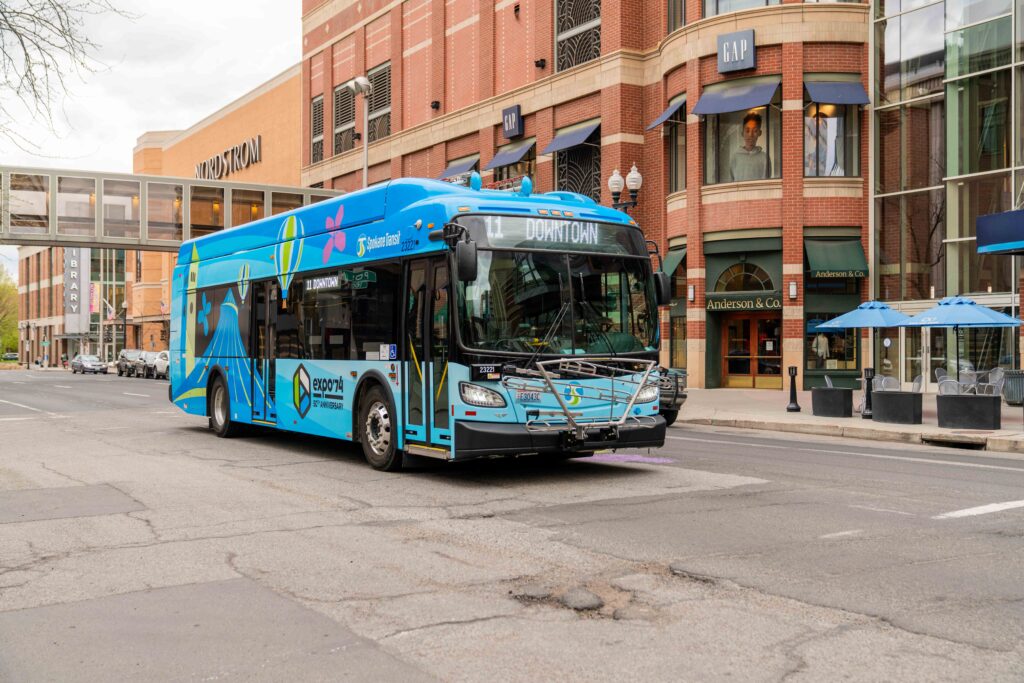 A city bus painted in bright blue with the text "Downtown" on the display is driving down a street lined with brick buildings. The buildings house various stores, including Gap and Anderson & Co. A pedestrian bridge connects two buildings above the street.
