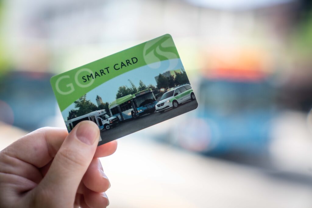 A hand is holding a green "SMART CARD" with images of various public transit vehicles, including buses and a train. The background is blurred, showing a street scene with indistinct buses and buildings.