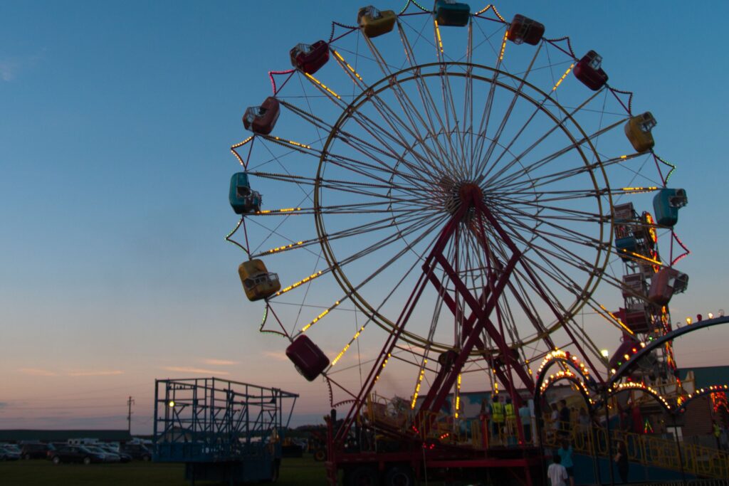 Ferris wheel at a fairground during sunset, with ride lights glowing and various fair attractions in the background.