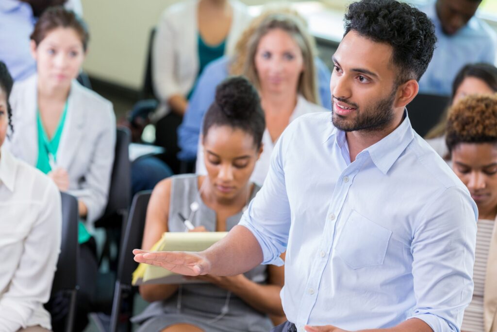 A man in a blue shirt speaks or asks a question during a group seminar or meeting; people in the background listen.