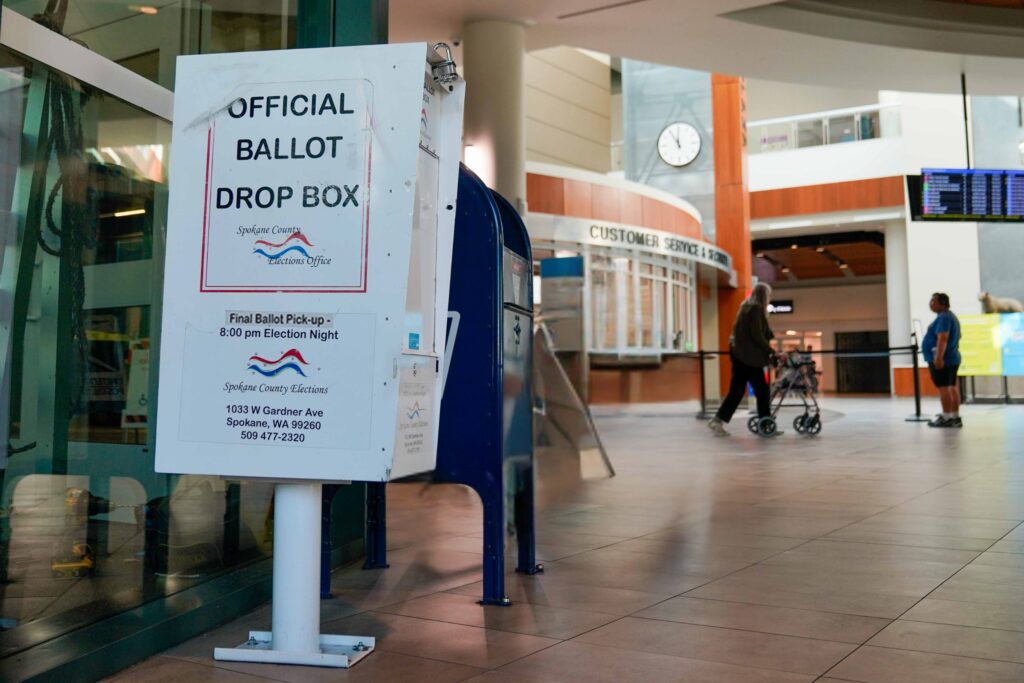 Official ballot drop box inside a building next to a glass door with two people walking in the background.