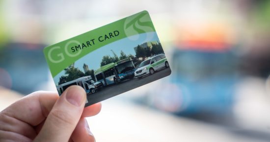 Close-up of a hand holding a transportation smart card. The card is green and blue, featuring images of various vehicles including a bus, a car, and a bicycle against a blurred outdoor background with indistinct colors.