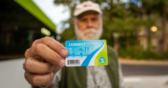 A bearded man wearing a cap holds a "CONNECT" public transit card towards the camera, displaying it prominently. The background shows blurred greenery and a bus, suggesting he is at a bus stop or in a transit area.