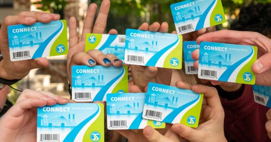 A group of people holding up multiple public transit cards with the word "CONNECT" on them. The cards feature a light blue and green design with a barcode and various icons. Some hands in the group are overlapping, and a leafy background is visible.