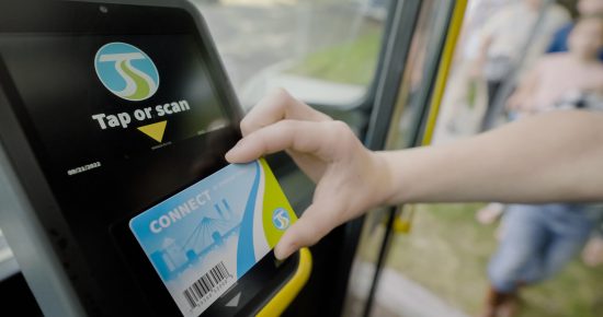A person scans a blue 'Connect' card on a card reader inside a public transportation vehicle. The screen of the reader displays an instruction to "Tap or scan." People are visible in the background.