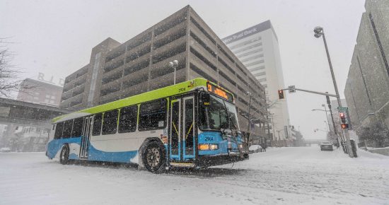 STA bus driving through heavy snowfall on a snowy Spokane street with buildings in the background.