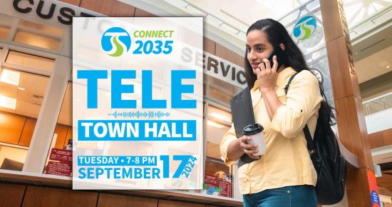 A woman holding coffee and a tablet talks on the phone next to a sign promoting a September 17 Tele Town Hall event.