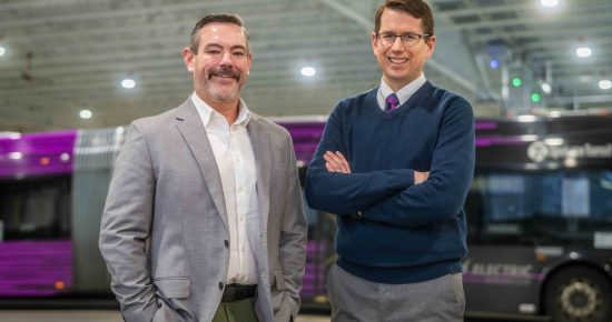 Two men standing in a bus depot, one in a suit and the other in a sweater, with purple buses in the background.